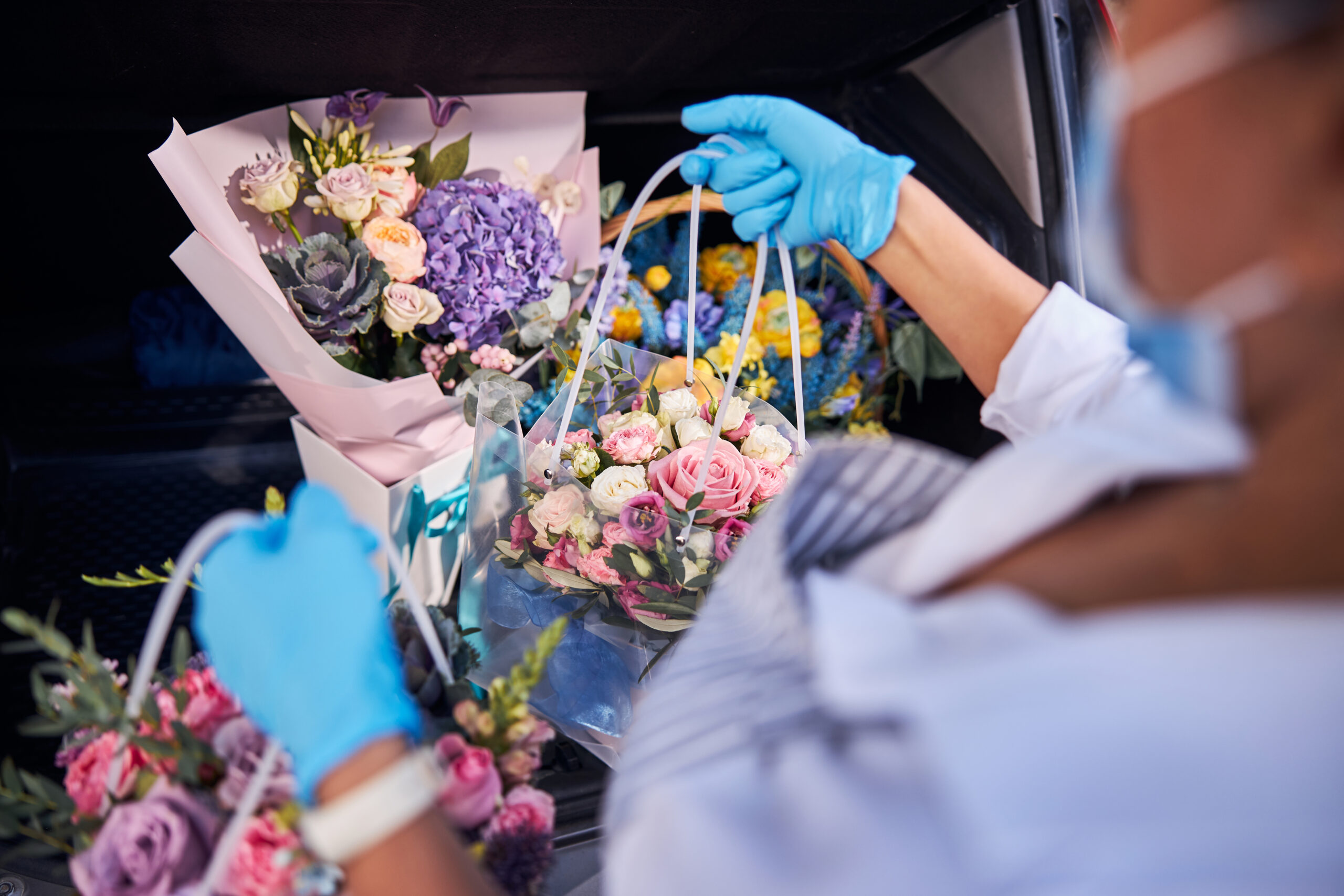Cropped photo of a mindful flower shop staff wearing rubber gloves while putting bouquets into a car trunk